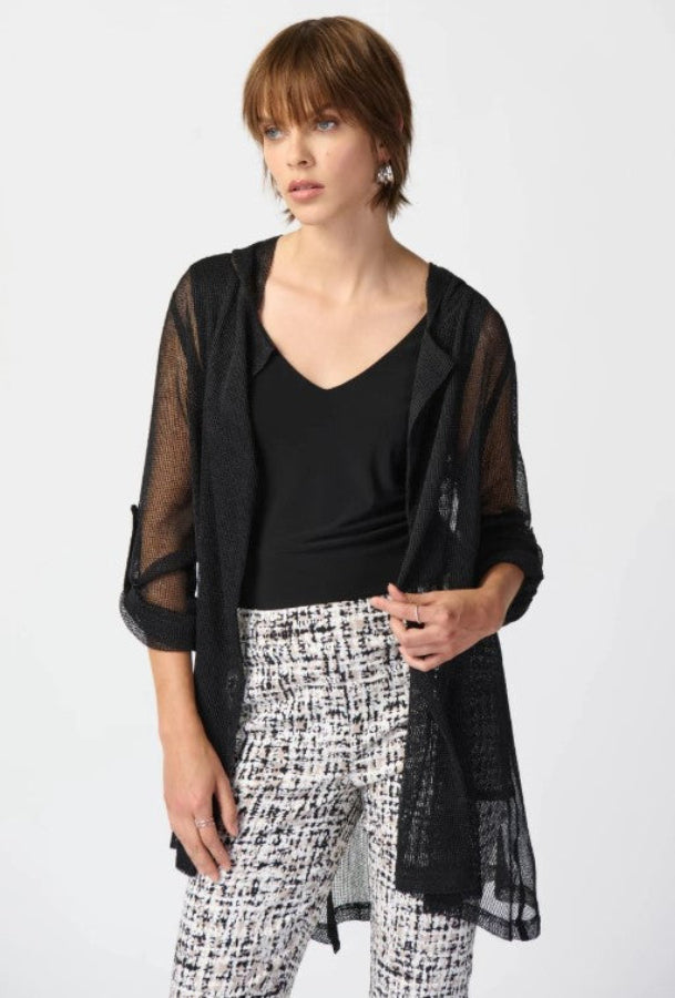 Woman wearing a black sheer jacket over a black top and patterned pants on a white background